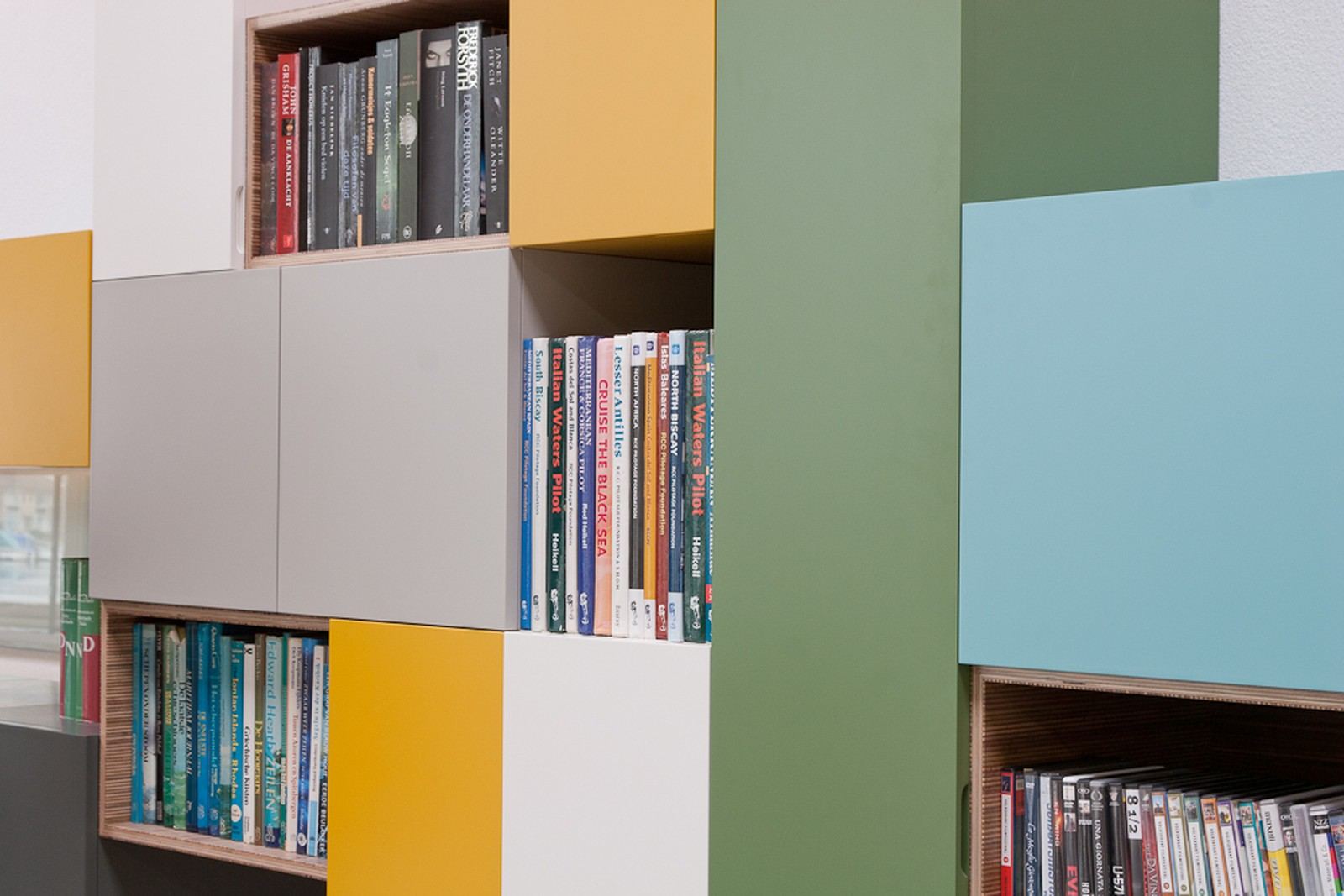 Detail van een open vak met dvd's en cd's, tussen groene, blauwgroene en witte kastblokken.