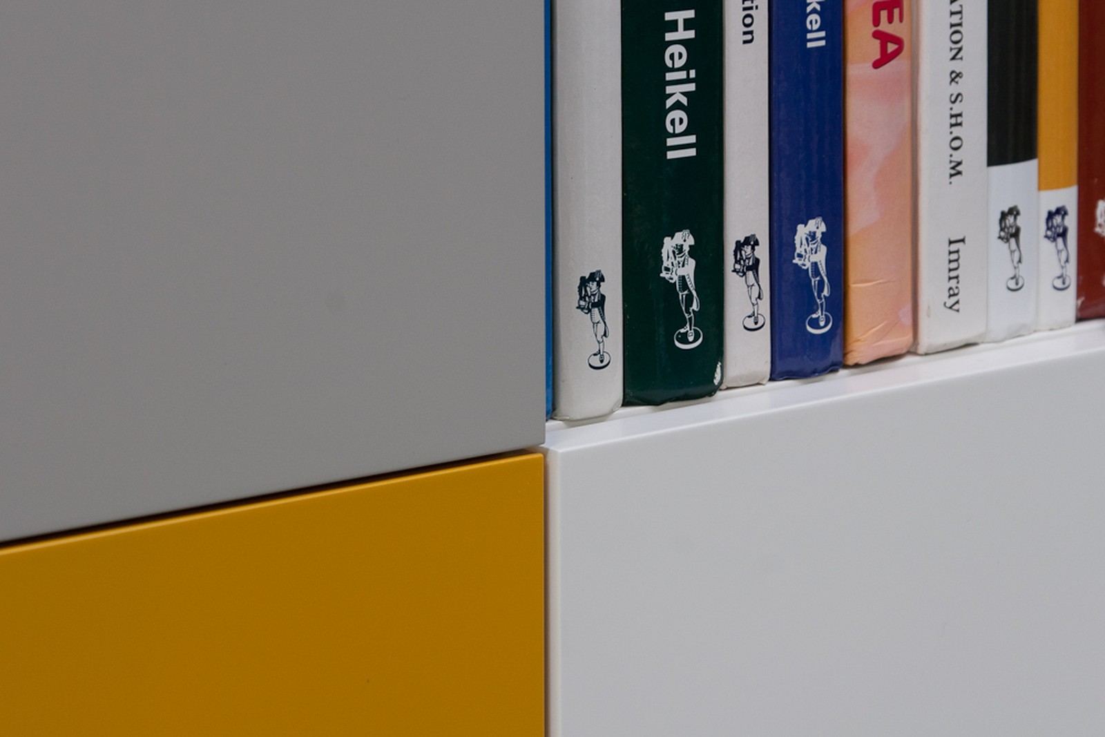 Detail van boekruggen in een vak tussen grijze en gele kastblokken.
