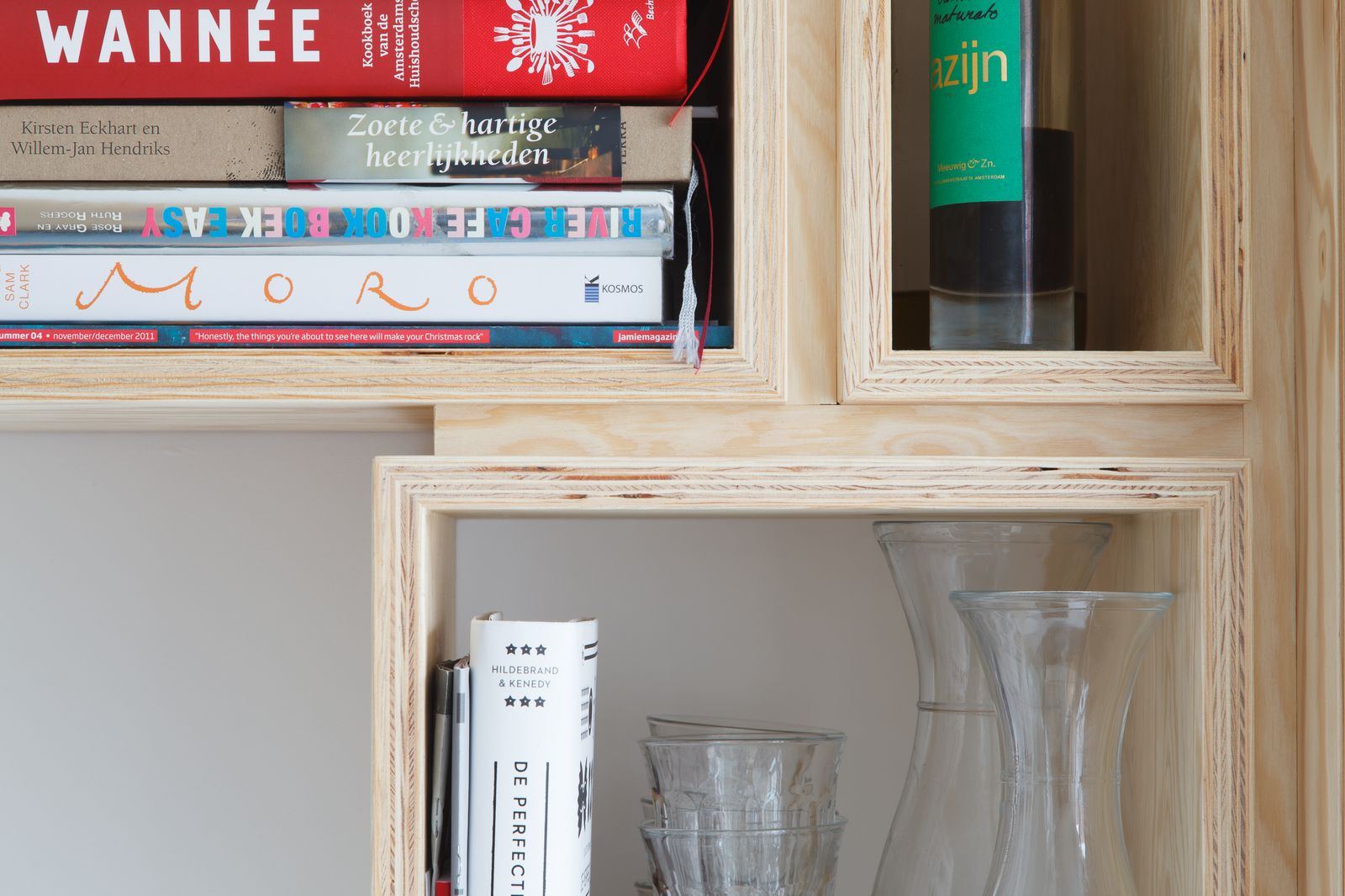 Open vakken van de wandkast met kookboeken, een fles en wijnglazen.