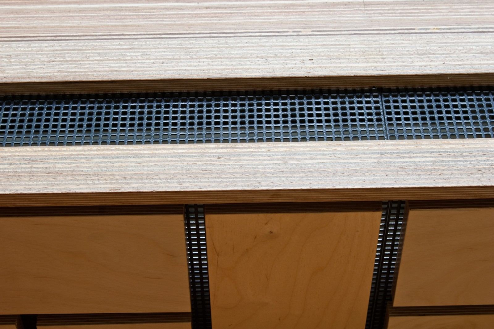 Detail van een geperforeerde metalen ventilatiestrook tussen de houten panelen van de bank.